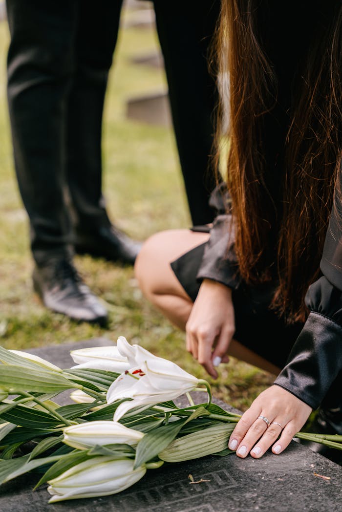 our-experience A woman kneels by a grave adorned with white lilies, symbolizing loss and remembrance.